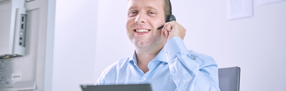 Mitarbeiter sitzt mit Headset vor dem Monitor und lächelt freundlich in die Kamera.