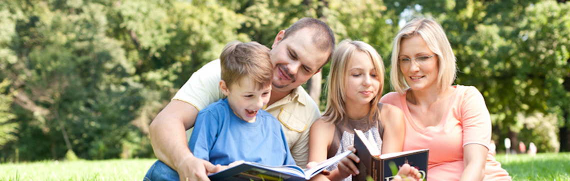 Eine Familie sitzt im Gras, die Eltern lesen ihren Kindern Geschichten vor.