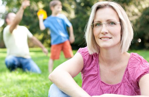 Eine Frau sitzt im Gras, hinter ihr spielt ihre Familie im Garten.